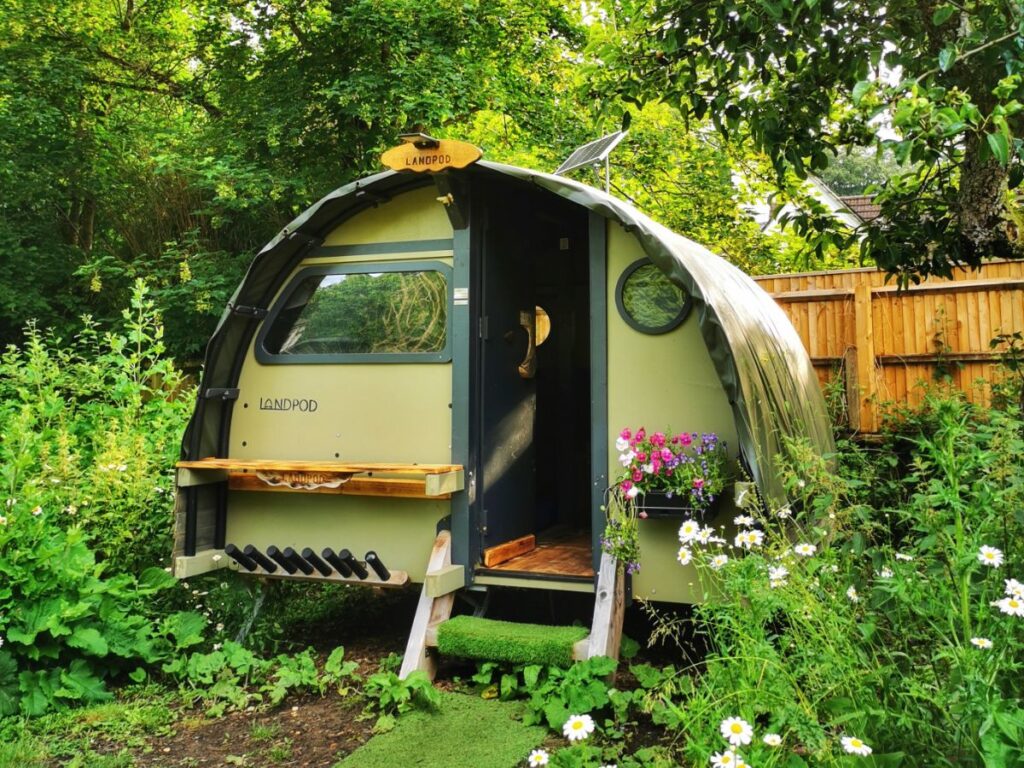 Eco-Friendly Land Pod - semi cylinder shaped pod with small windows and flowers in front - Glamping Short Breaks in the UK