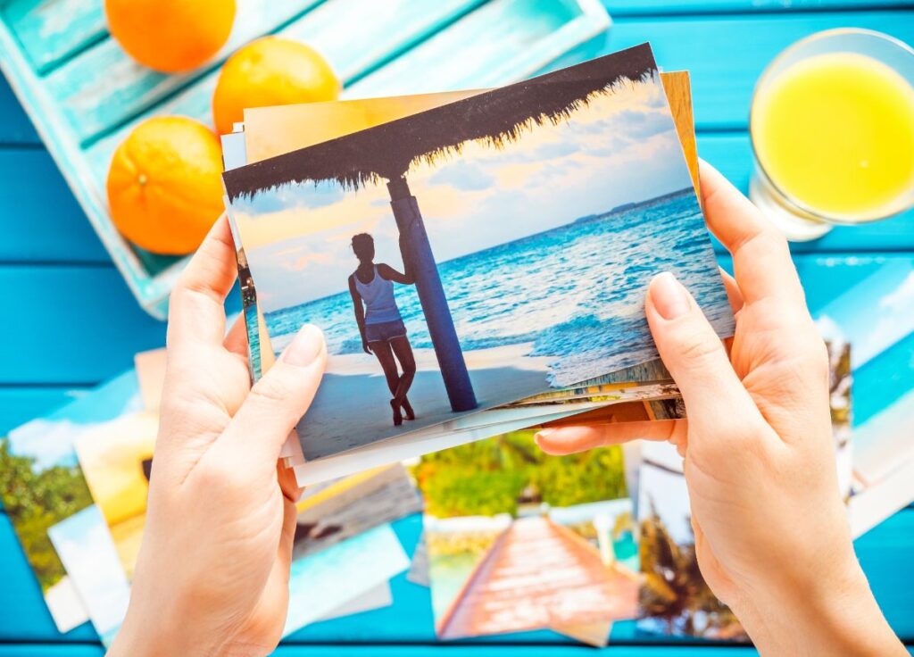 Hands holding a photograph of a woman by the sea, with bright blue table background with more photos and a couple of oranges on the table -How to Preserve Travel Memories