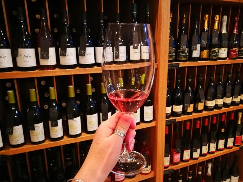 Sampling Some Delicious Czech Wine - My hand holding a glass of rose wine with shelves of bottles behind