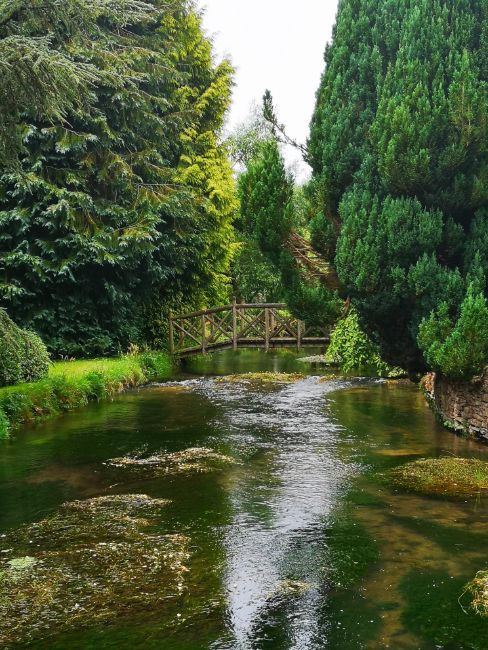 Picuresque River with trees on either side and a wooden footrbidge in the distance - The River Windrush provides a Quiet Refuge from the Crowds