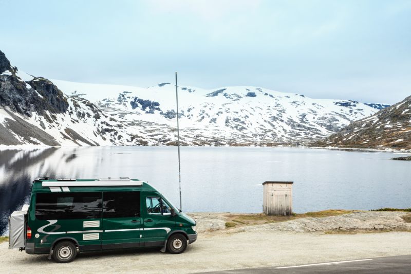 Green campervan in front of a cold lake in Norway with snow-covered hills beyond - Cold Weather Brings Extra Challenges for Campervan Road Trips