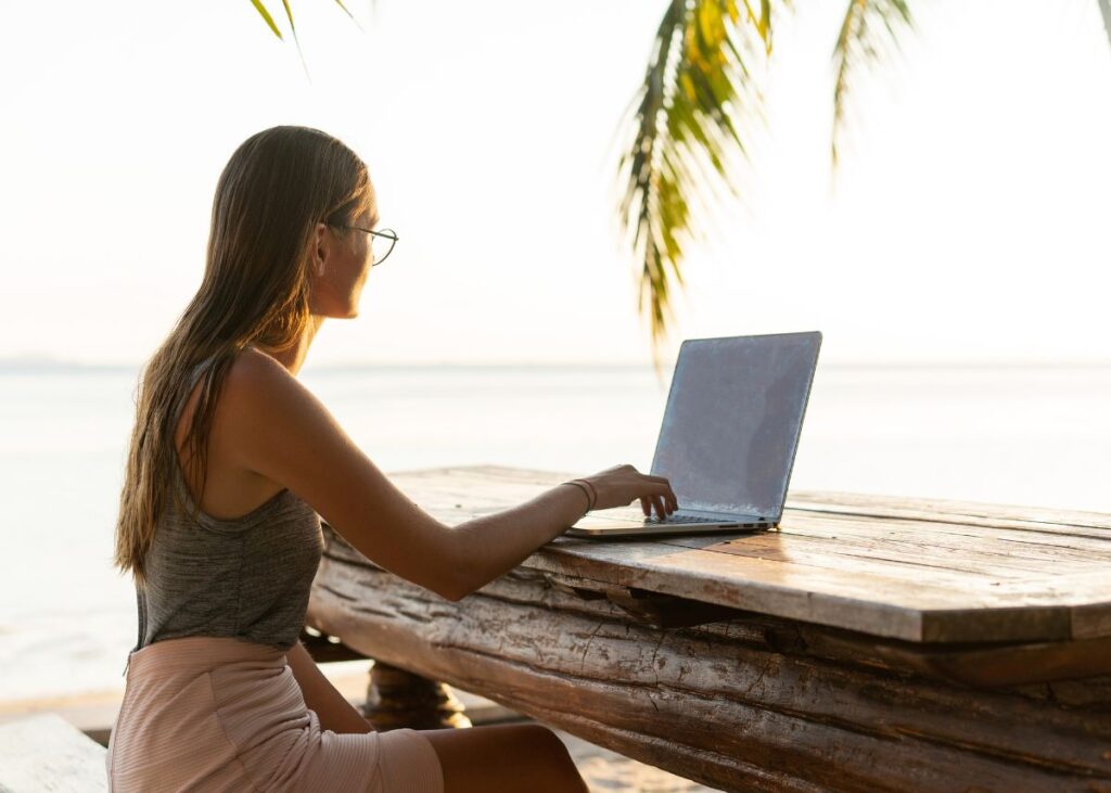 Woman sitting on a bench by the sea on her laptop - Digital Nomad Lifestyle Tips