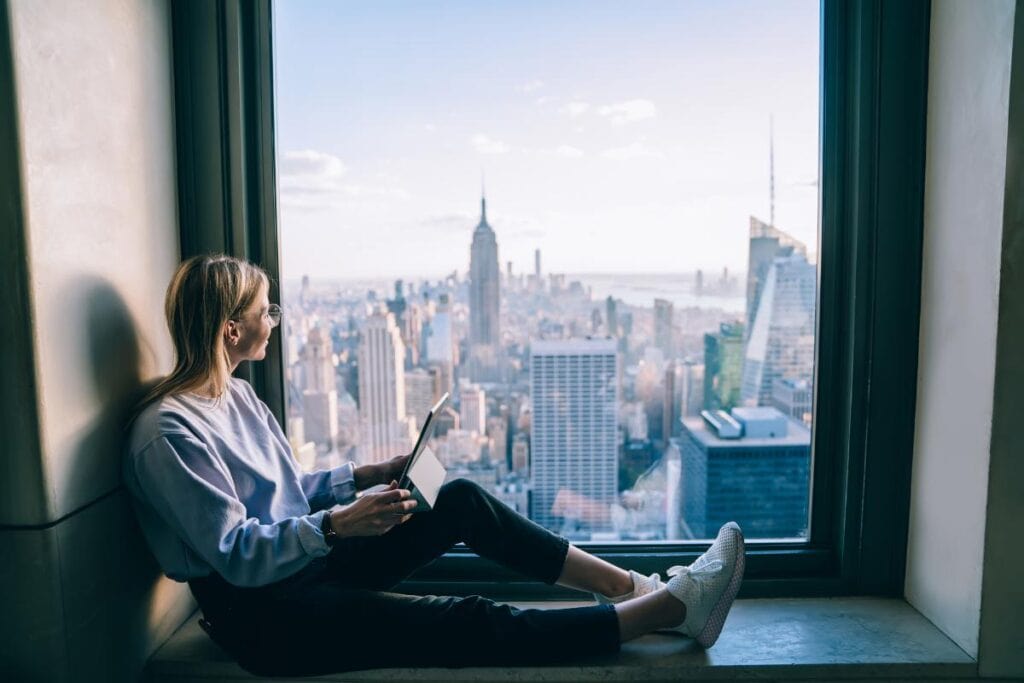 Woman Working on a Laptop at a Window with a View of New York - Digital Nomad Loneliness